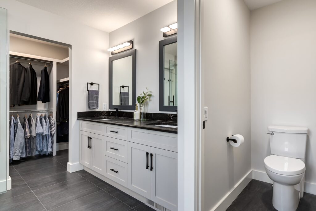 Modern bathroom with double sinks, black countertops, two mirrors, and a toilet on the right. There’s a walk-in closet with clothes visible through an open doorway on the left. Walls are light gray and floors are dark tile.