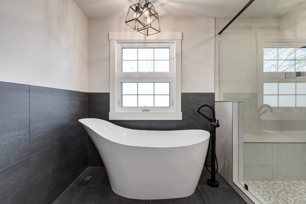 Modern bathroom with a white freestanding bathtub, black floor-mounted faucet, dark gray tile walls, large window, glass shower enclosure, and geometric ceiling light fixture.