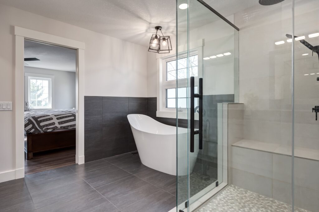 Modern bathroom with large glass shower, freestanding white bathtub, gray tile floor and walls, and a window providing natural light. An open doorway leads to a bedroom with a bed and two windows.
