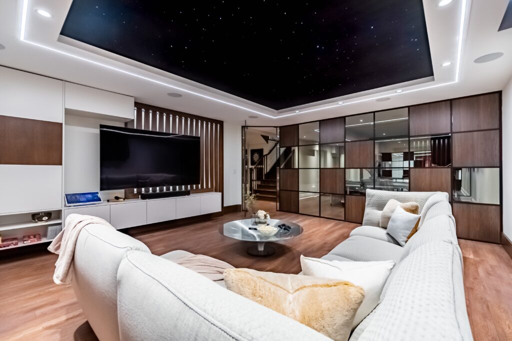 Modern living room with a large sectional sofa, coffee table, wall-mounted TV, wood and glass panel wall, built-in shelves, and a ceiling designed to look like a night sky with star-like lights.