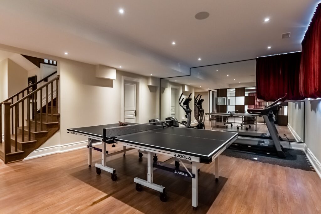 A home gym with wood floors features a ping pong table, treadmill, elliptical machine, and mirrored wall. Stairs lead to another floor, and red curtains partially cover one wall. Recessed lighting brightens the room.