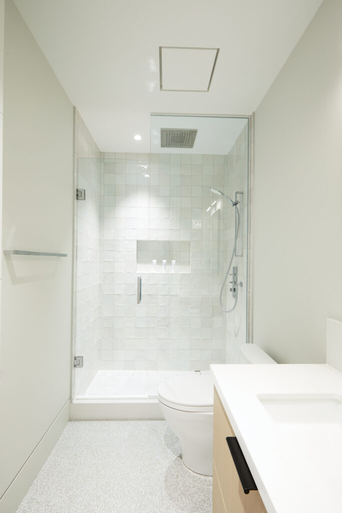 A modern, minimalist bathroom with a glass-enclosed shower, white textured tiles, a wall-mounted showerhead, a toilet, and a white sink with a light wood vanity. The space is bright and clean.