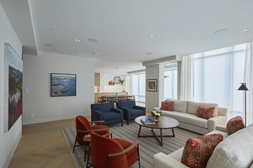 A modern living room with large windows, cream-colored sofas, two blue armchairs, two red chairs, a round wooden coffee table, patterned cushions, wall art, and an open view into the dining area and kitchen.