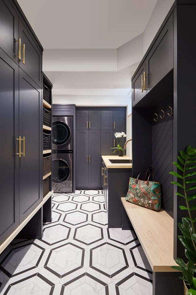 Modern laundry and mudroom with dark cabinetry, gold handles, a geometric black-and-white tile floor, stacked washer and dryer, a bench with a colorful handbag, and a potted plant in the corner.