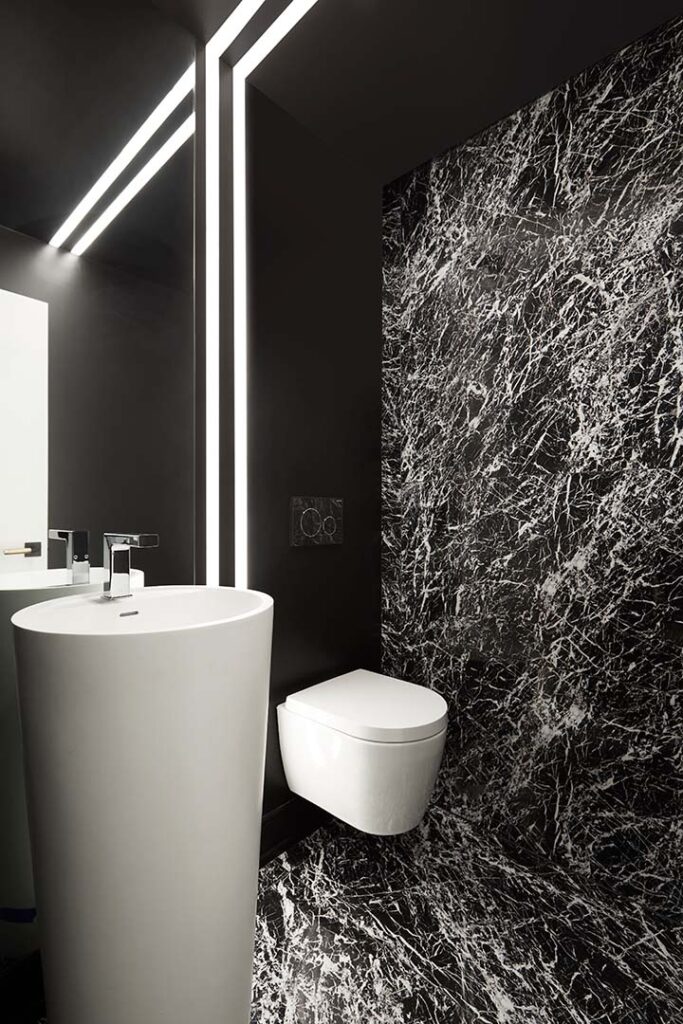 Modern bathroom with a cylindrical white sink, wall-mounted white toilet, and black marble walls and floor with white veining. Vertical LED lights line the corner, creating a sleek, contemporary look.