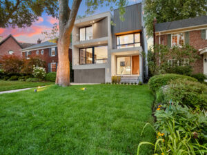 Modern two-story house with large windows and wood accents, set on a lush green lawn with trees and garden beds, surrounded by traditional brick homes at sunset.