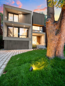 Modern two-story house with large windows, balconies, and a spacious lawn. A tree stands in the foreground, and a pathway leads to the entrance. The sunset sky adds warm colors in the background.