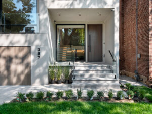 Modern house entrance with glass door, gray steps, and minimal landscaping. The address number 387 is on the left wall. There are small bushes in front and potted plants near the steps. The facade is light-colored and sleek.