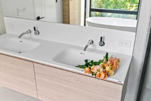 A modern bathroom with a double sink and light wood cabinets. One sink holds a bouquet of peach and yellow roses. A large mirror and window with green trees outside are visible in the background.