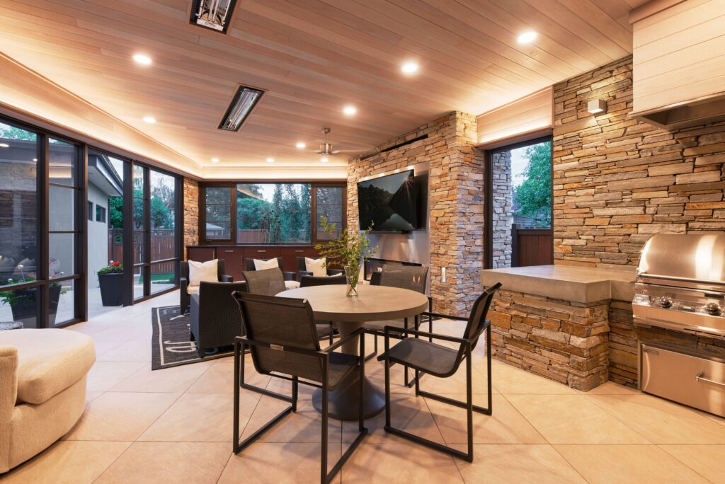Modern covered patio with stone walls, round dining table with four black chairs, built-in stainless steel grill, and large windows overlooking a yard. Warm lighting creates a cozy, inviting atmosphere.