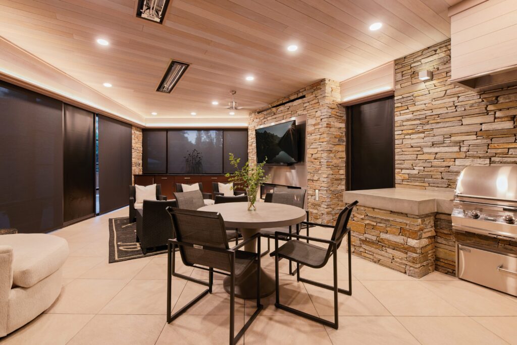 Modern indoor patio with stone walls, a round table and four chairs, a gas grill, flat-screen TV, and large windows with black shades. Warm lighting creates a cozy, inviting atmosphere.