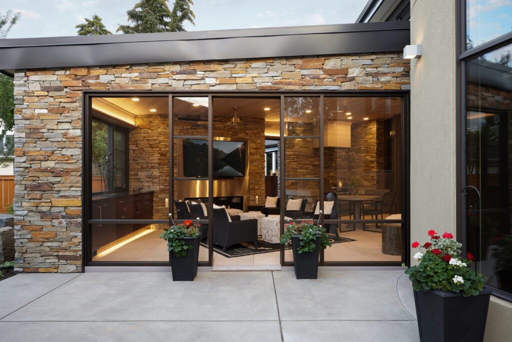 A modern patio with glass sliding doors opens to a cozy, stone-walled living area. Two black planters with red and white flowers sit on the concrete patio outside. Warm lights illuminate the interior.
