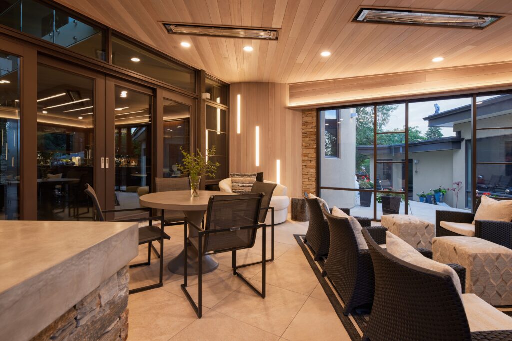 Modern indoor-outdoor living space with a round dining table and chairs, cozy lounge seating, large windows, wood ceiling, accent lighting, and views of a patio area and surrounding greenery.