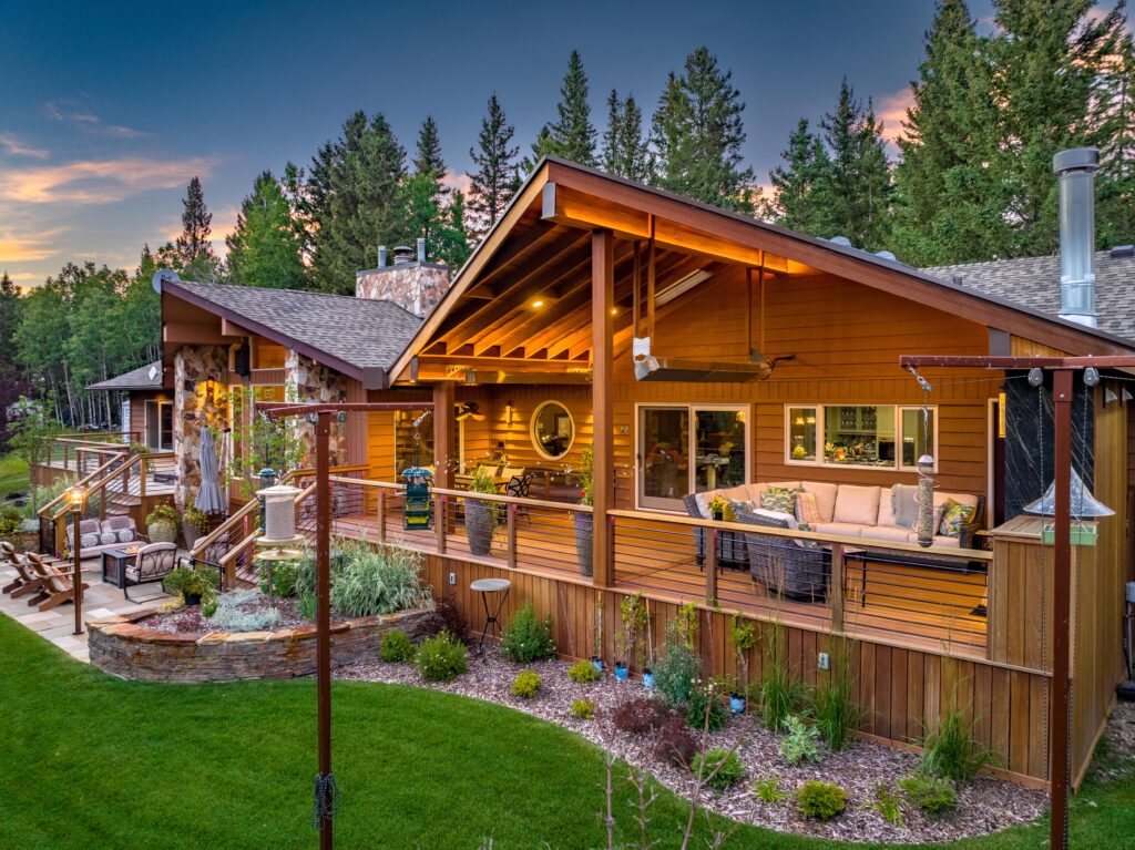 A spacious wooden house with a large covered deck, outdoor seating areas, and landscaped garden, surrounded by tall trees at sunset. The warm lighting creates a cozy and inviting atmosphere.