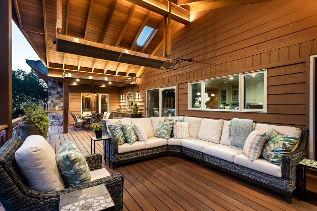 Spacious wooden patio with a beige cushioned sectional sofa and decorative pillows, wicker armchair, dining table, and warm overhead lighting beneath a sloped roof with exposed beams. Large windows line the house wall.