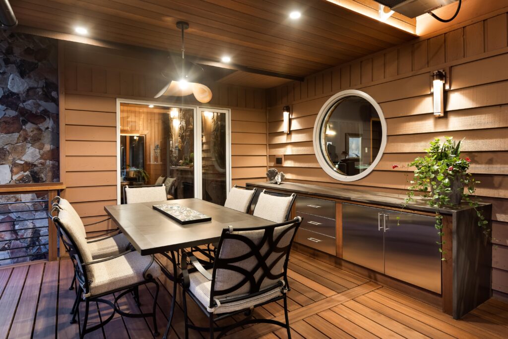 A cozy, modern outdoor patio with wood paneling, a rectangular dining table with six cushioned chairs, a ceiling fan, built-in cabinets, warm lighting, and a round window above a countertop with a potted plant.