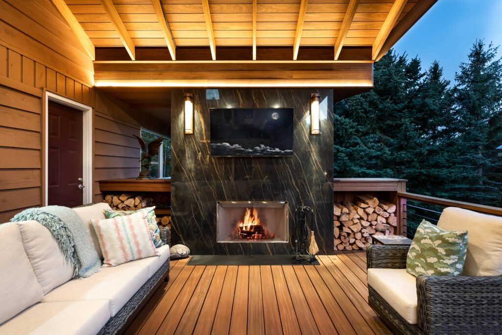 A cozy outdoor patio with cushioned seating, a wooden deck, a modern black fireplace with a TV above it, stacked firewood on both sides, and trees visible in the background at dusk.