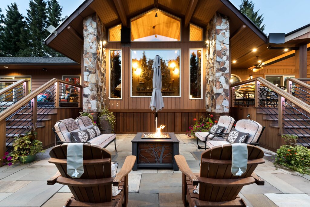 A cozy outdoor patio with wooden chairs and cushioned sofas surrounds a fire pit. The space is framed by a modern wooden house with large windows, stone columns, and string lights, with trees visible in the background.