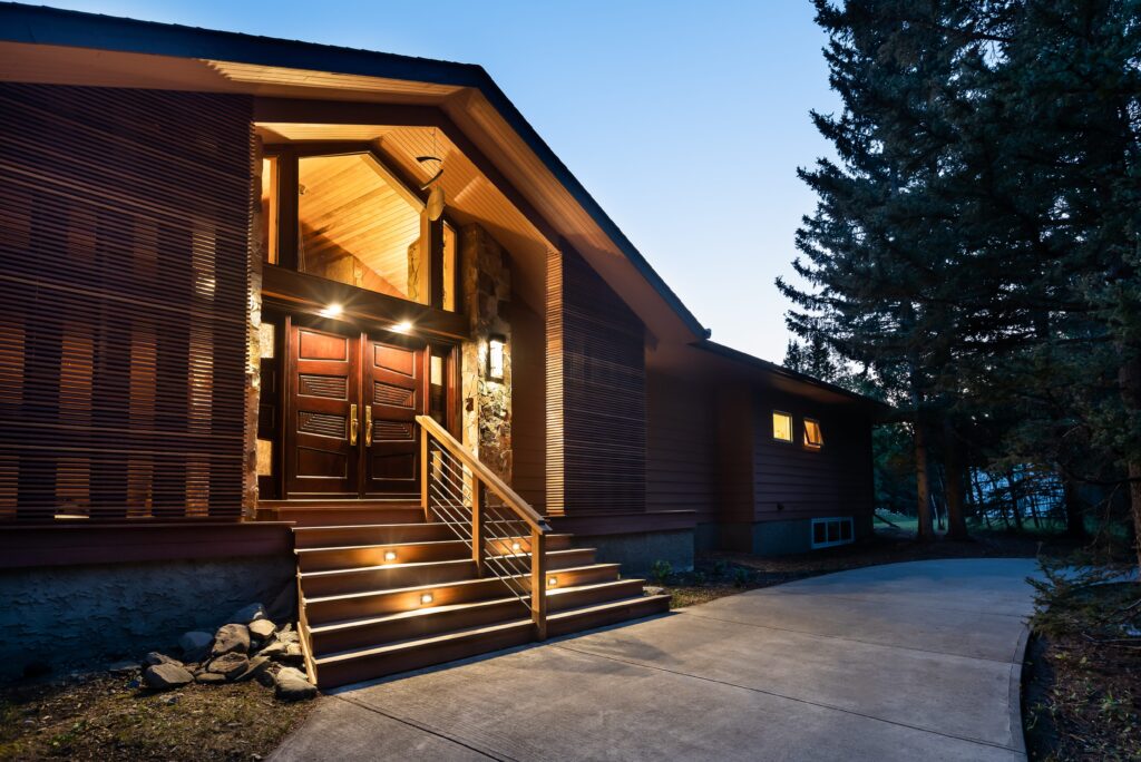 Modern house exterior at dusk with lights illuminating wide front steps leading to a wooden double door entrance; the walkway curves around landscaped rocks and large trees frame the right side of the image.
