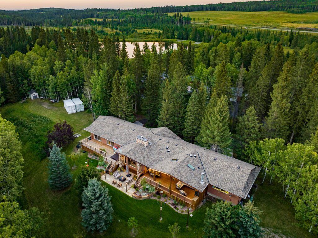 Aerial view of a large house surrounded by dense green trees, open lawn, and a small pond in the background, set in a lush, forested landscape.