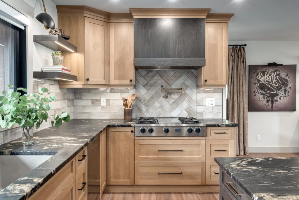 Modern kitchen with light wood cabinets, black marbled countertops, a built-in gas stove, chevron-patterned tile backsplash, overhead range hood, and decorative plants and books on shelves.