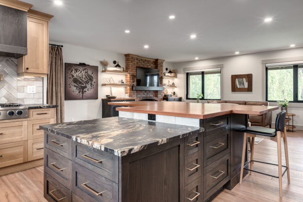 Modern kitchen with two-tone islands, dark lower cabinets, light upper cabinets, a black marble countertop, bar stools, wood flooring, and an open view into a cozy living area with a brick fireplace and TV.
