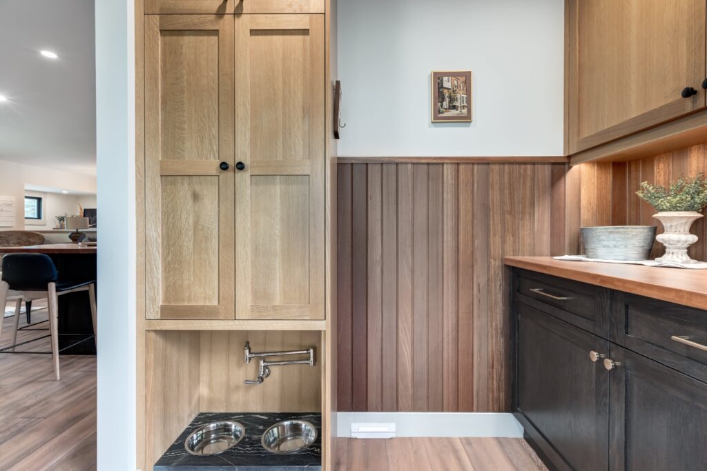 A modern kitchen features light wood cabinets, dark lower drawers, a wood-paneled wall, and a built-in pet feeding station with two metal bowls beneath a tall cabinet. A plant and a framed picture add decorative touches.