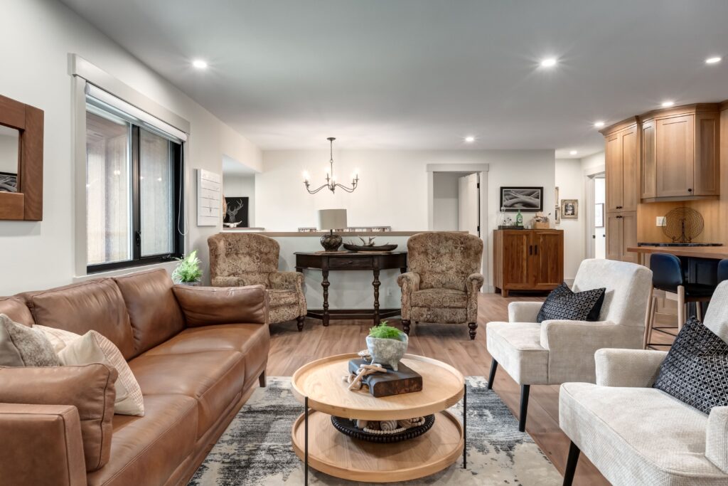 A cozy living room with a brown leather sofa, two beige armchairs, and two patterned chairs. A round wooden coffee table sits on a geometric rug. In the background is a dining area with a chandelier and wooden furniture.
