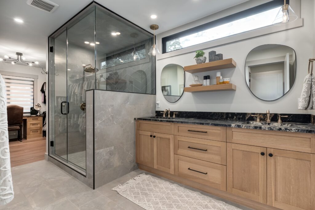 Modern bathroom with a glass shower, double vanity with black countertops, wooden cabinets, two round mirrors, wall shelves with decor, and a white textured rug on a gray tile floor.