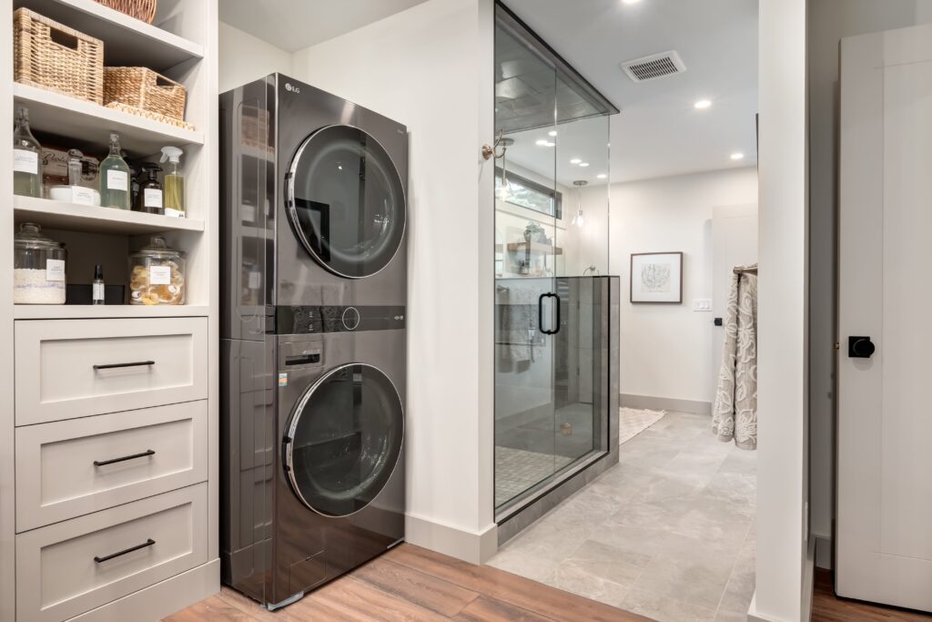 A modern laundry room with a stacked washer and dryer next to built-in shelves and drawers, adjacent to a glass-walled shower in a spacious, well-lit bathroom.