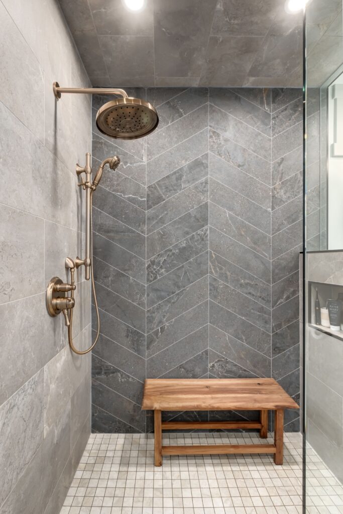 Modern shower with gray tile walls in a chevron pattern, a brass rain showerhead and handle, a wooden bench, and a small shelf with toiletries. The floor is made of square light tiles.