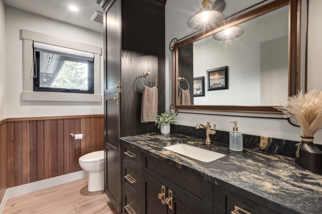 A modern bathroom with a black marble countertop, dark wood cabinets, a rectangular mirror, a wall-mounted light, and a small window above a toilet. Decorative plants and framed art accents the space.