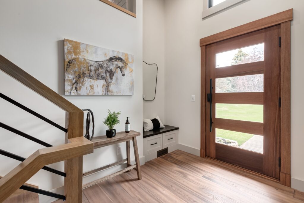 A modern entryway with a wooden front door, a small bench with drawers, a wall mirror, a table with a plant and decor items, and a painting of a horse on the wall next to a staircase.