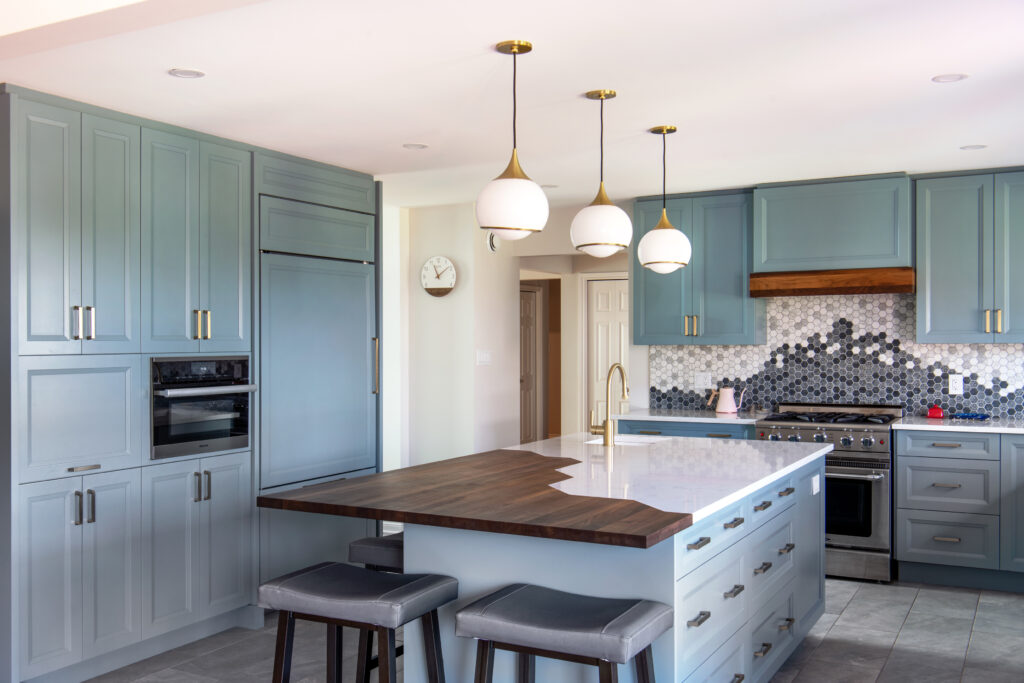 Modern kitchen with blue-gray cabinets, a large island with a wood and white countertop, pendant lights, gray stools, and a backsplash featuring hexagonal tiles in various shades of gray and white.