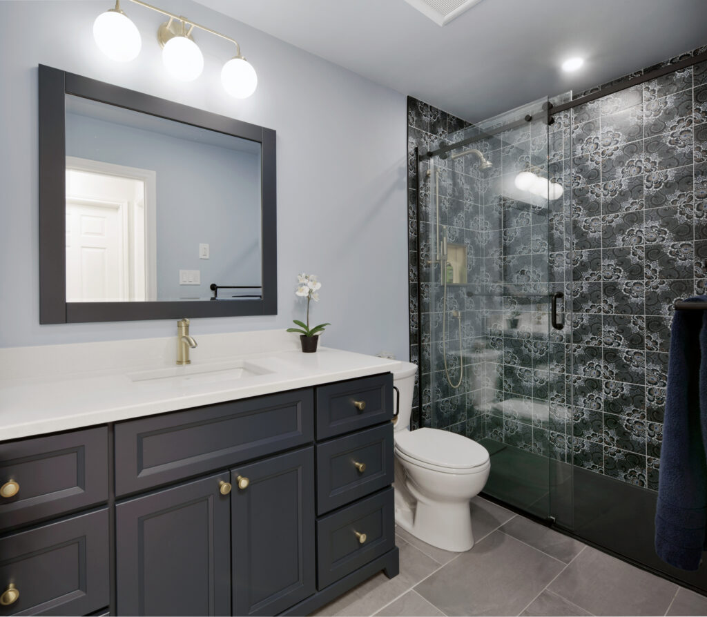Modern bathroom with a navy vanity, white countertop, brass fixtures, a large mirror, and a potted orchid. The shower features a glass door and decorative black-and-white tiles. Gray floor tiles complete the space.