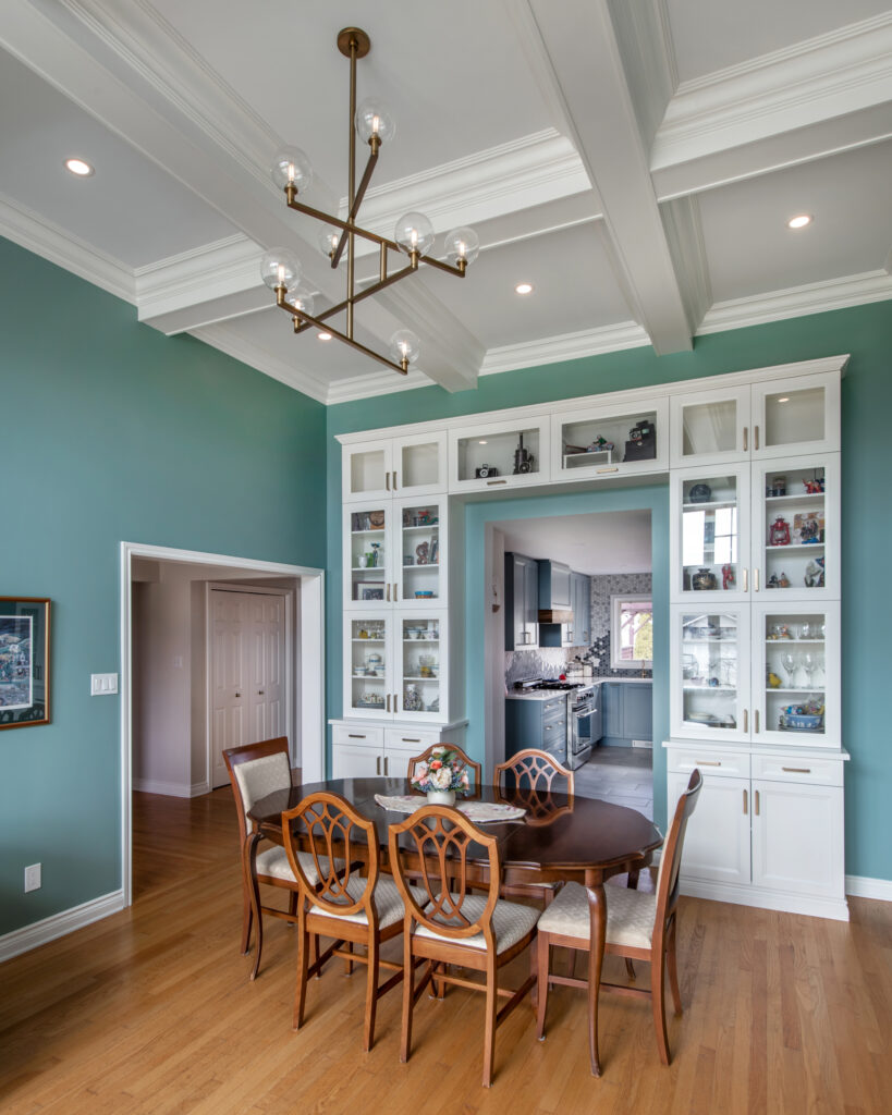 A dining room with teal walls, wooden floor, a round wooden table with six chairs, a modern chandelier, and white built-in cabinets displaying decor. The room opens to a kitchen in the background.