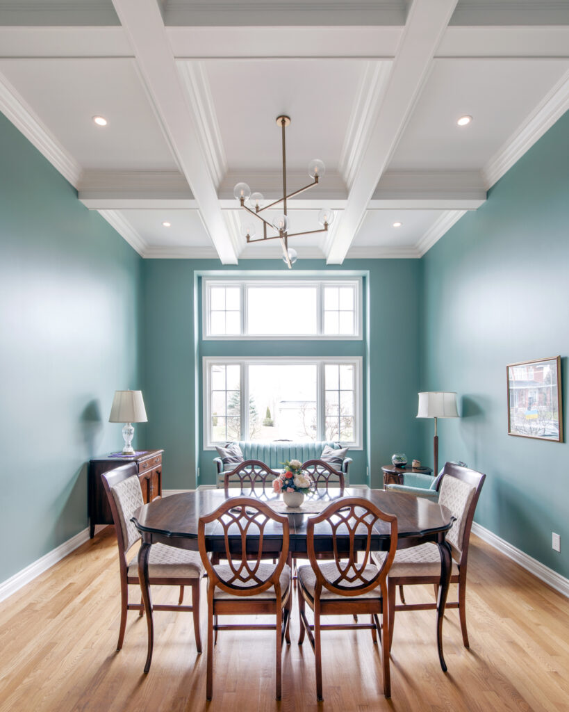 A bright dining room with teal walls, a wooden table and six chairs, large windows, hardwood floors, two side tables with lamps, and a modern chandelier hanging from a coffered ceiling.