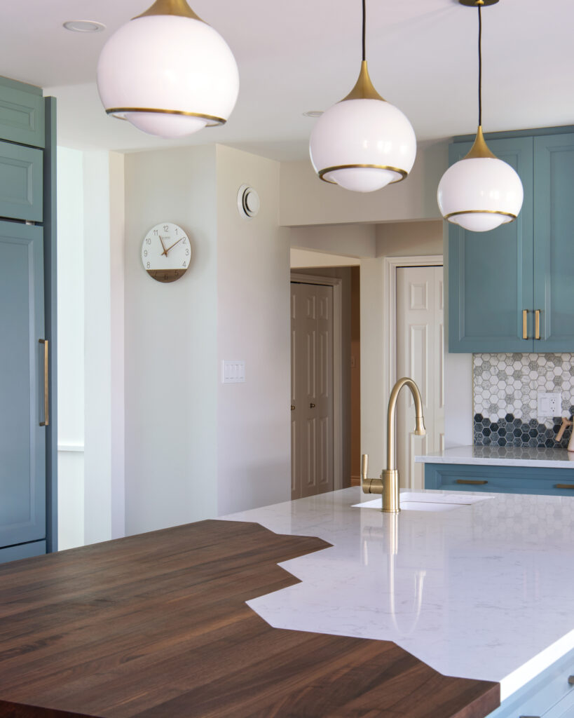 Modern kitchen with blue cabinets, hexagonal tile backsplash, brass fixtures, three globe pendant lights, and a unique island countertop design featuring both wood and white marble surfaces. A white wall clock is visible in the background.