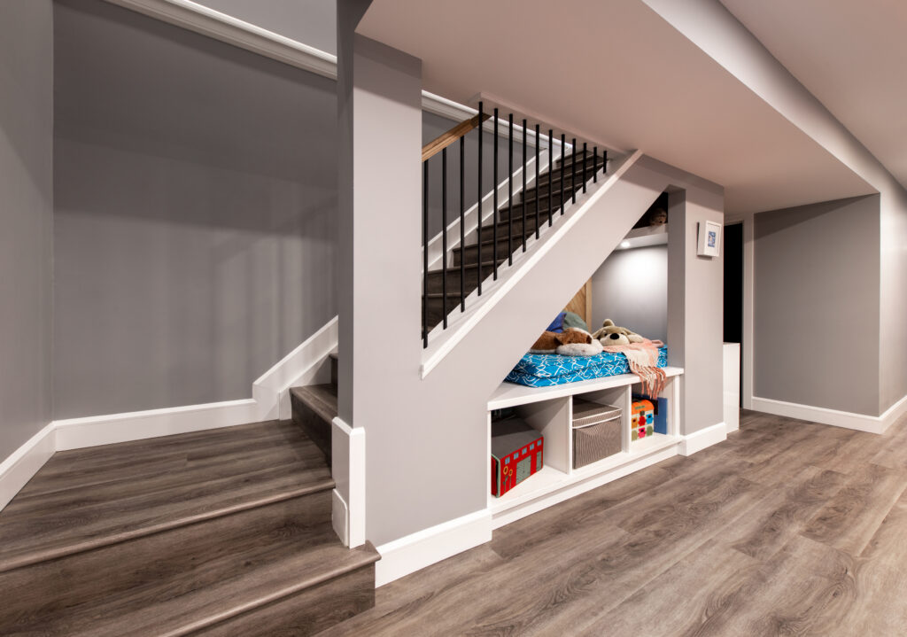 A modern basement features gray walls, wood flooring, and a cozy nook with a bed and toys built under a staircase with black railings. The area is neatly organized and well-lit.