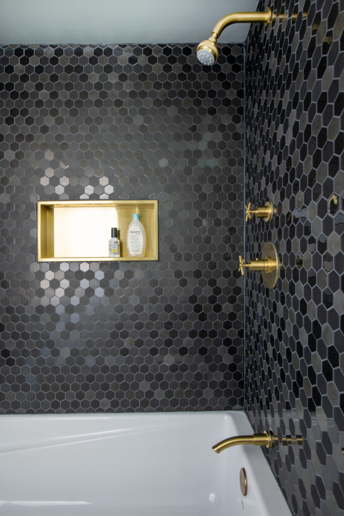 Shower with dark hexagonal tile walls, gold fixtures, and a built-in recessed shelf holding toiletries above a white bathtub.