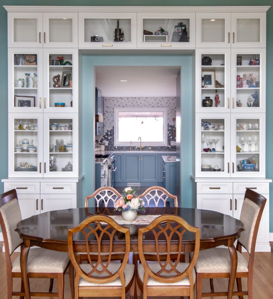 A dining room with a dark wooden table and six chairs, surrounded by white display cabinets filled with decor. An open doorway reveals a blue kitchen with a sink and window at the back.