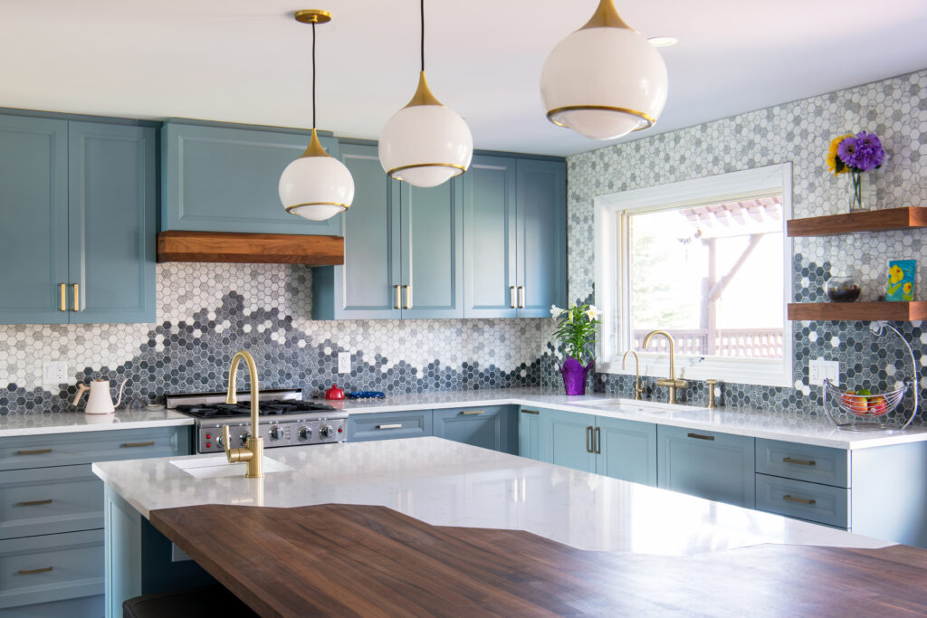 Modern kitchen with blue cabinets, gold fixtures, white and wood island, three pendant lights, a hexagon tile backsplash in shades of gray, and a window bringing in natural light. Flowers and decor sit on open shelves.