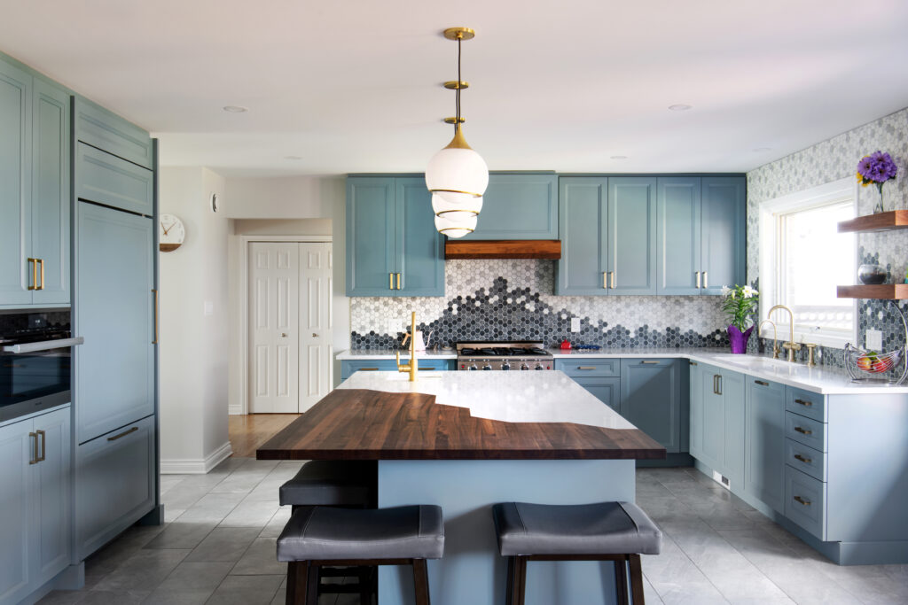 Modern kitchen with light blue cabinets, a large island with a wooden countertop, two gray stools, and geometric tile backsplash. Pendant lights hang above the island; a window lets in natural light.