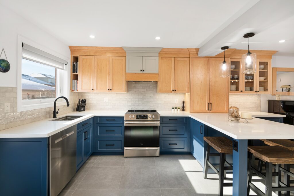 Modern kitchen with natural wood upper cabinets, blue lower cabinets, stainless steel oven, white countertops, large island with bar stools, pendant lights, and a window overlooking a snowy landscape.