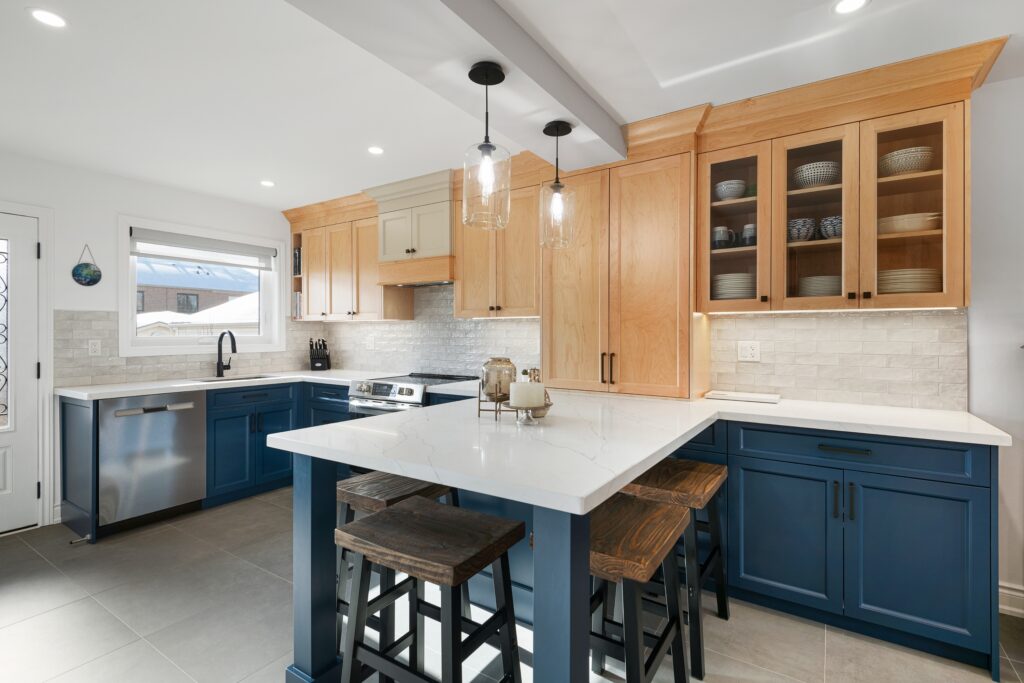 Modern kitchen with light wood upper cabinets, blue lower cabinets and island, white quartz countertops, stainless steel appliances, two pendant lights, and bar stools around the island.