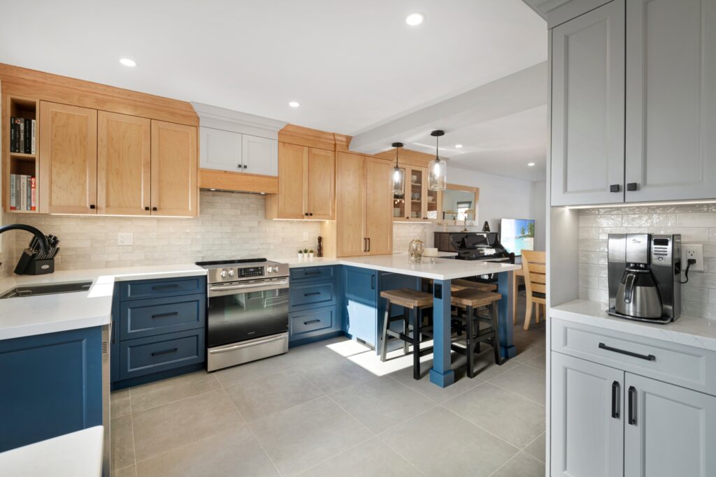 Modern kitchen with blue and wood cabinets, stainless steel stove, white countertops, pendant lights over a small island with two chairs, and a coffee maker on a side counter. Light gray tile floor and subway tile backsplash.