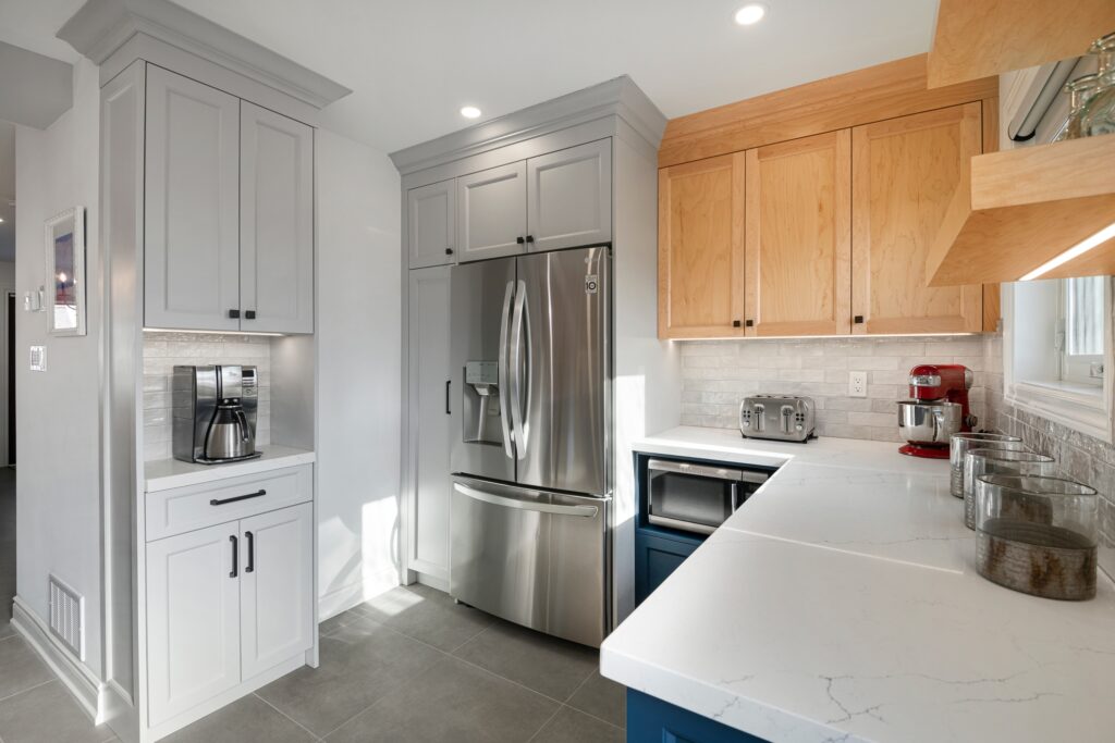 Modern kitchen with stainless steel refrigerator, white and wood cabinets, coffee maker, toaster, microwave, and mixer on white countertops, under bright lighting with gray floor tiles and subway tile backsplash.