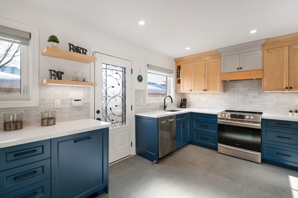 Modern kitchen with blue lower cabinets, light wood upper cabinets, stainless steel appliances, white countertops, gray tile floor, open shelves, and a decorative glass door, brightly lit by recessed lighting and natural light.