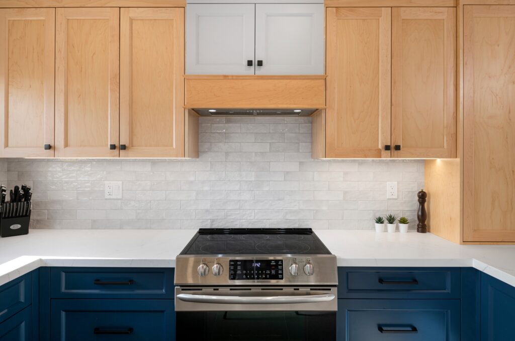 Modern kitchen with light wood upper cabinets, blue lower cabinets, a stainless steel electric stove, white countertop, white subway tile backsplash, knife block, pepper mill, and small potted plants.