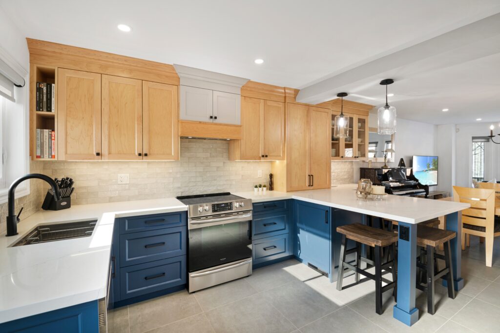 Modern kitchen with light wood upper cabinets, blue lower cabinets, stainless steel appliances, white countertops, a tiled backsplash, two pendant lights, and a breakfast bar with two wooden stools.